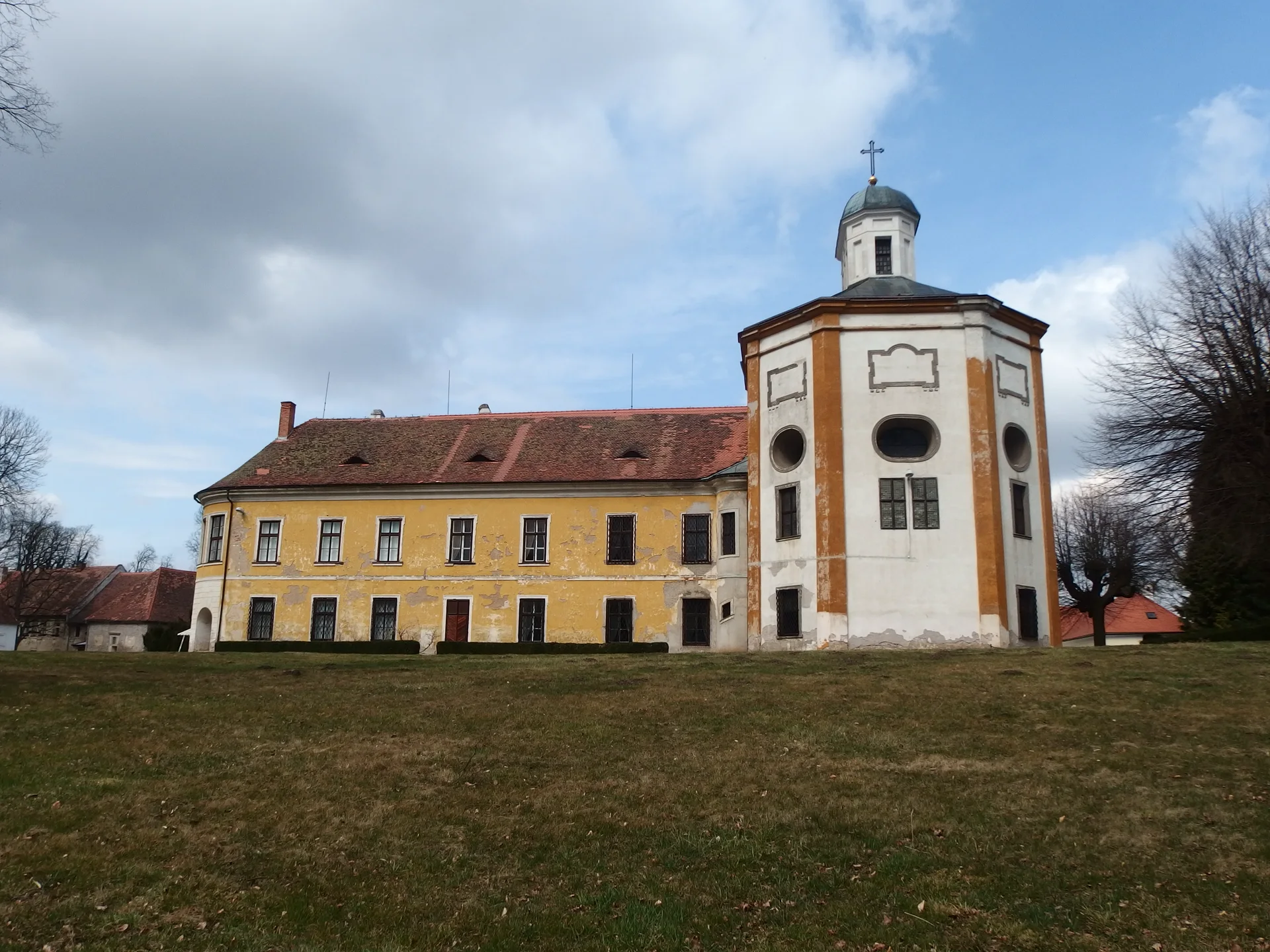 Zámek Choltice od louky v parku, žluté křídlo a věž zámecké kaple pod oblačným nebem