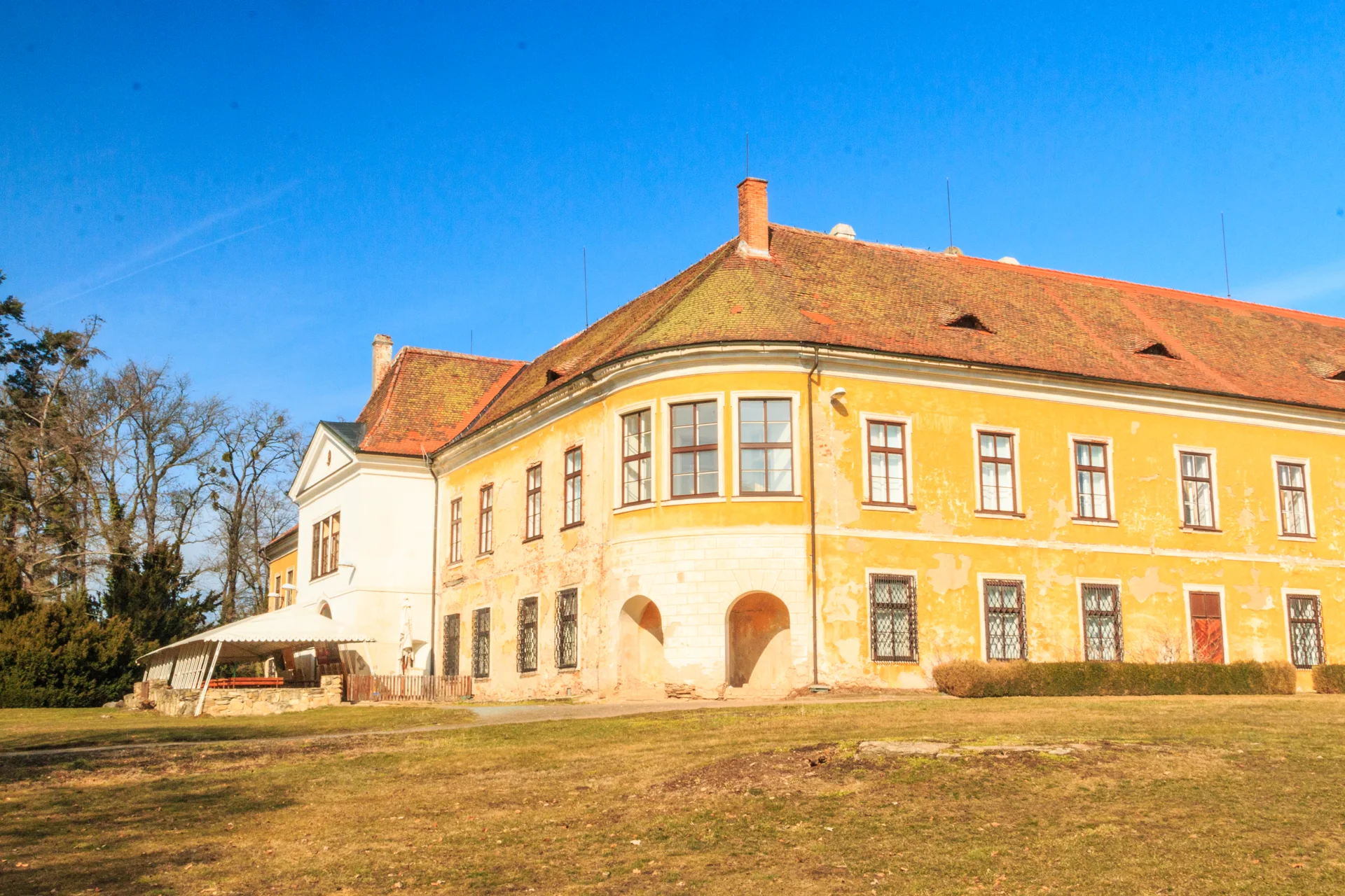 Žlutý zámek Choltice s červenou střechou a půlkruhovým rizalitem, před ním trávník za slunného dne