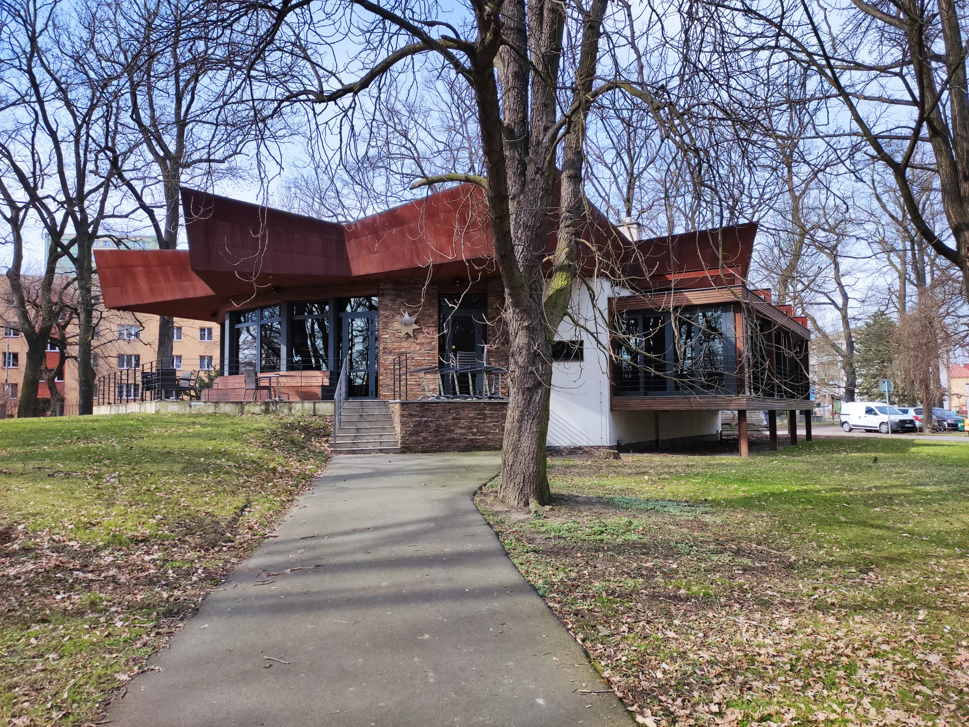 Restaurace Hvězda s červenou vlnitou střechou na kraji parku, chodník mezi stromy