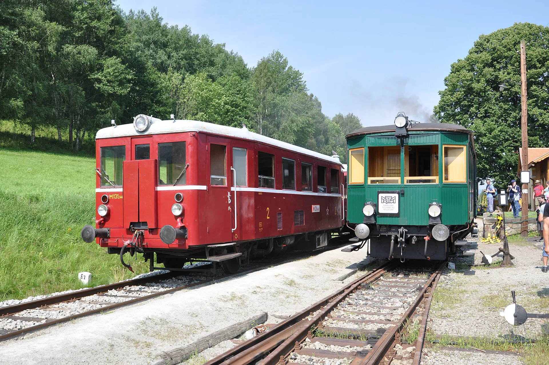 Dva historické motorové vozy, červený a zelený, stojící na kolejích u stanice
