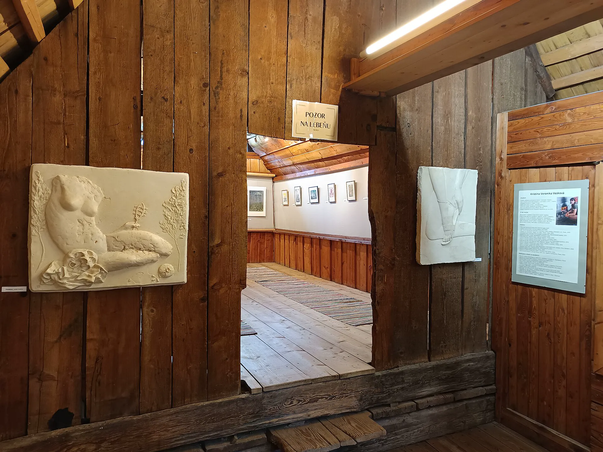 Interiér expozice v roubené dřevěnici s reliéfem a průhledem do další místnosti
