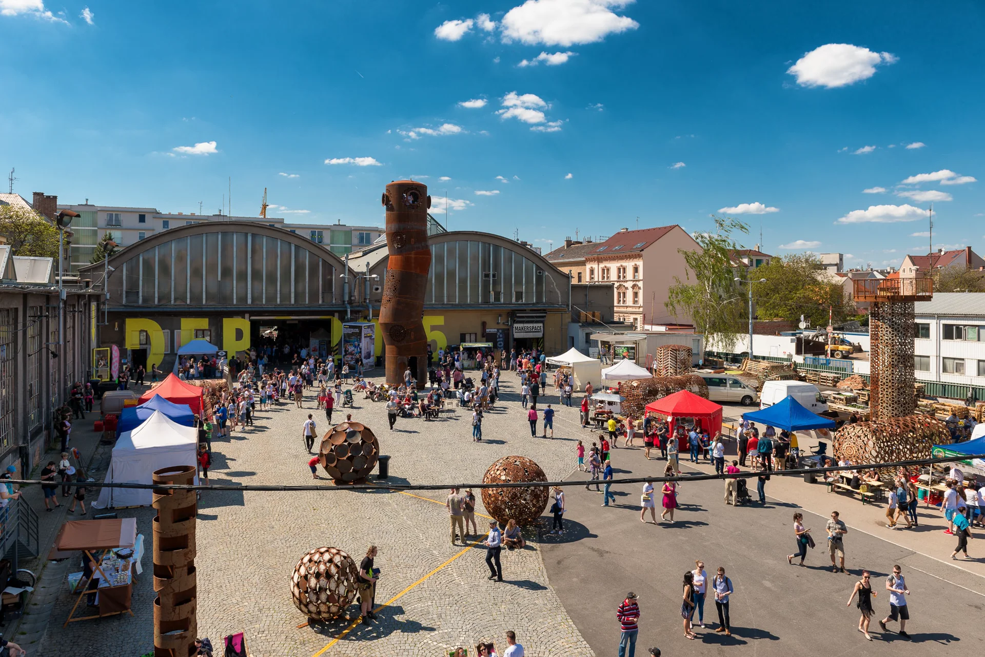 Letecký pohled na areál DEPO2015 s halami, rezavým komínem a stánky na nádvoří