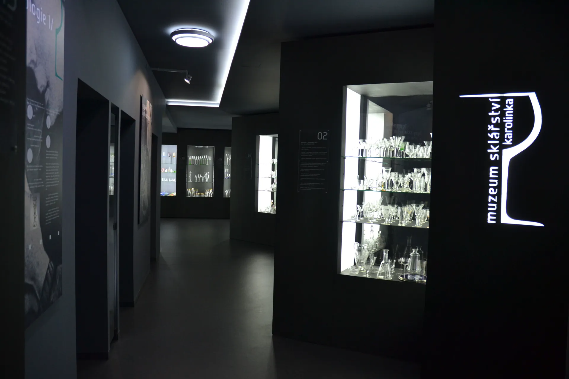 Tmavá chodba expozice s prosvětlenými vitrínami skla a svítícím logem muzea