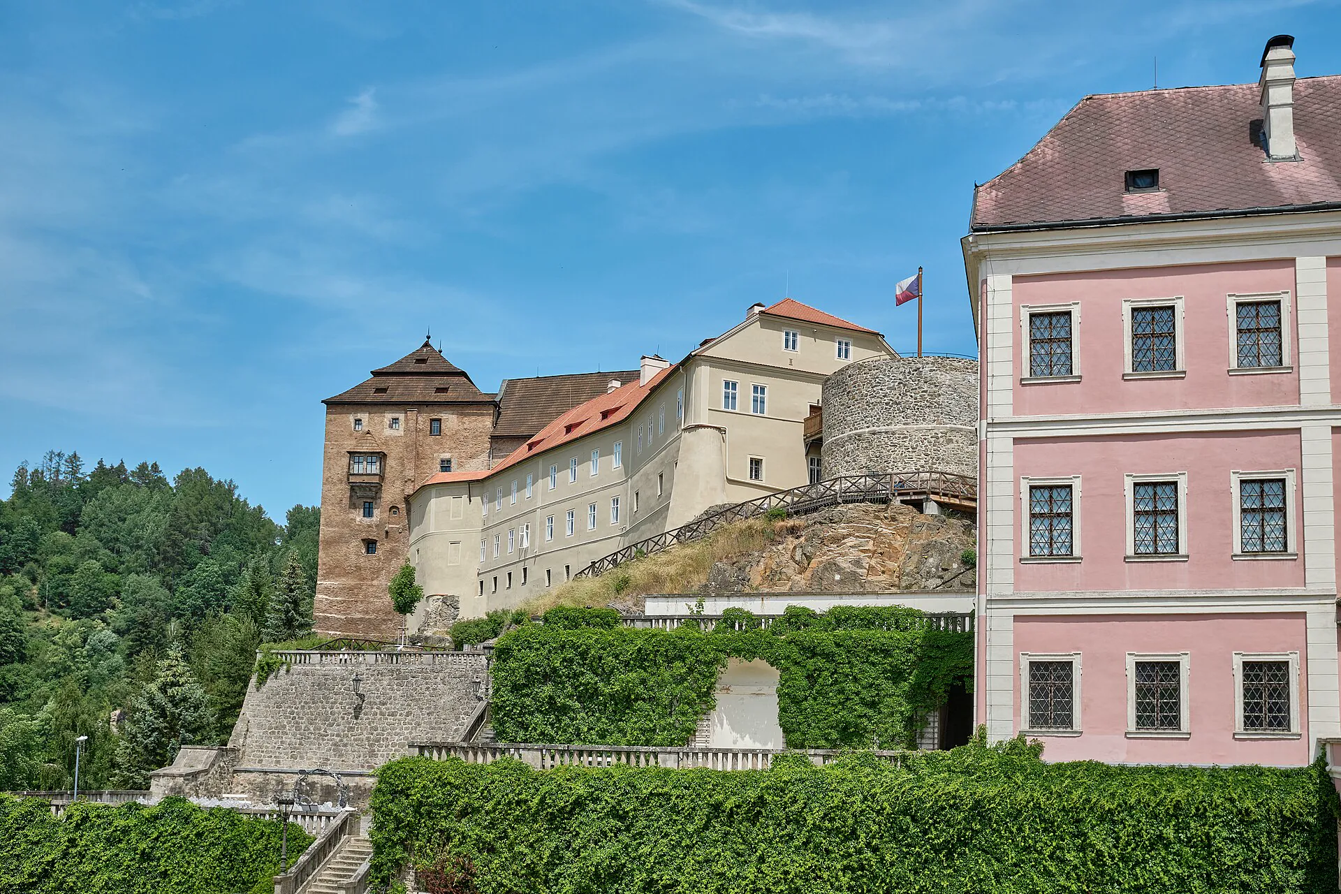 Gotický hrad s věží nad terasami a barevnými domy podhradí